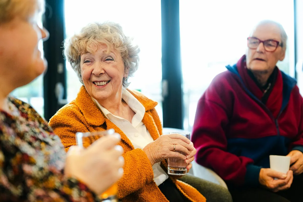 Ältere Frau lacht im Gespräch mit anderen bei einem Getränk